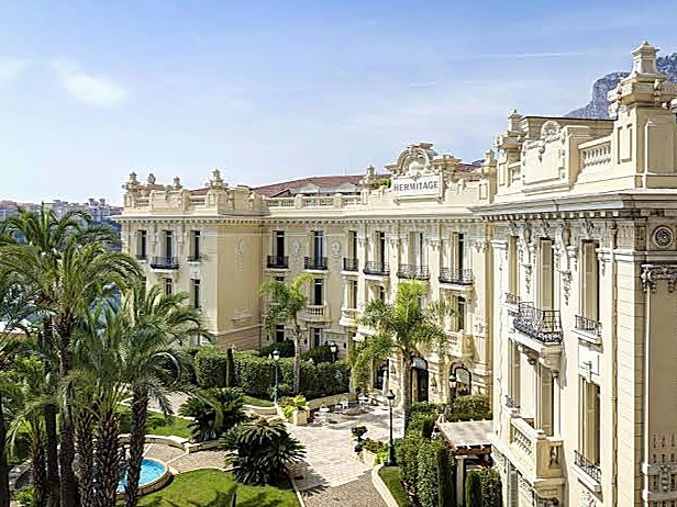 Fachada de piedra clara con balcones de hierro, balaustradas y frontón coronado, sobre un jardín de palmeras, con el peñón detrás.