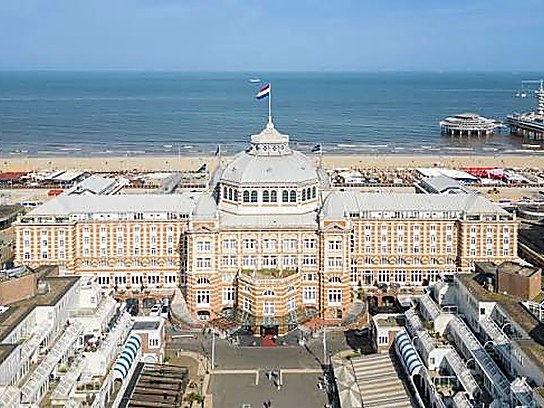 Vista aérea de un edificio de ladrillo y piedra con cúpula central y pabellones laterales, entre la playa del mar del Norte y el frente urbano.